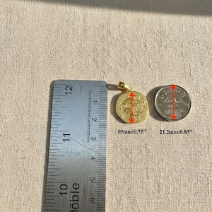 Puede incluir: Un colgante dorado con un dise&ntilde;o circular, que mide 19 mm de di&aacute;metro. El colgante se muestra junto a una regla y una moneda para comparar el tama&ntilde;o. El colgante tiene un peque&ntilde;o lazo para una cadena.