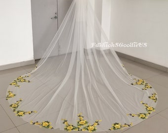 May include: A long white bridal veil with a delicate floral pattern in yellow. The veil is made of soft tulle and has a scalloped edge.