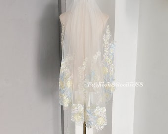 May include: A white bridal veil with delicate floral embroidery in shades of blue and yellow. The veil is made of soft tulle and has a scalloped edge.