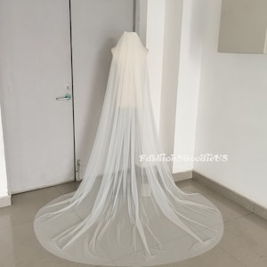 May include: A long, white tulle wedding veil with a simple design. The veil is draped over a mannequin and is shown from the back.