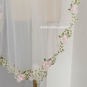 May include: A white tulle veil with a delicate floral embroidery in pink and green. The embroidery is around the edge of the veil.