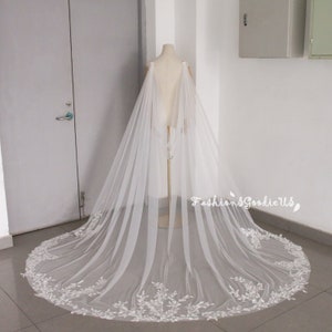 May include: A white tulle wedding veil with a delicate floral lace trim. The veil is long and flowing, with a train that extends to the floor. The veil is designed to be worn over a wedding dress.
