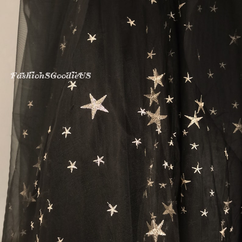 Op de afbeelding: Zwarte tule stof met zilveren glittersterren. De stof is doorschijnend en heeft een zachte, vloeiende textuur.  FashionSGoodieUS is op de stof gedrukt.