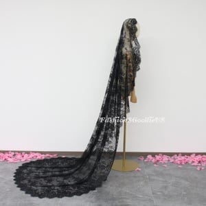 May include: A long black lace veil with a scalloped edge. The veil is draped over a mannequin and is shown from the side.
