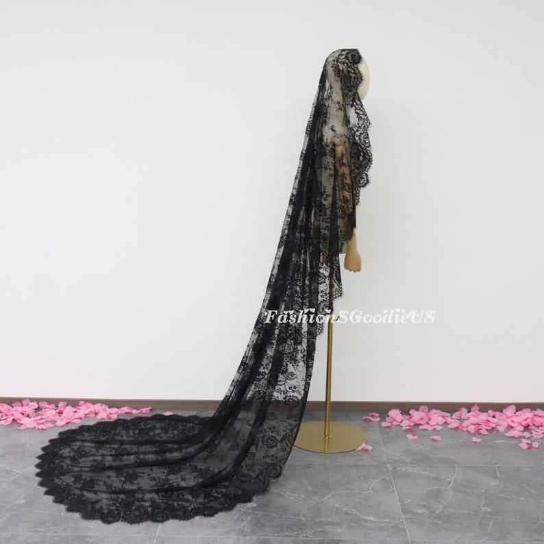 May include: A long black lace veil with a scalloped edge. The veil is draped over a mannequin and is shown from the side.