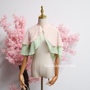 May include: A layered cape in pastel colors, displayed on a mannequin. The top layer is a soft pink, with a mint green layer underneath. The cape is lightweight and appears to be made of chiffon. The background features pink cherry blossom branches.
