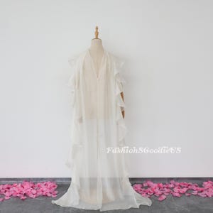 Pu&ograve; includere: Un abito maxi bianco in chiffon con maniche a volant e scollo a V. L'abito &egrave; su un manichino e ha un aspetto fluido ed etereo. Il testo "FashionSGoodieUS" &egrave; visibile nell'angolo in basso a destra dell'immagine.
