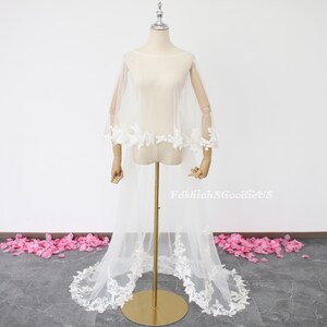 May include: White tulle bridal cape with delicate lace trim and floral embroidery. The cape has a long train and is displayed on a mannequin.