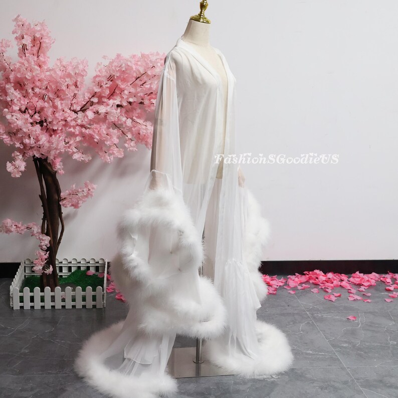 May include: A white, sheer robe with feather detailing on the sleeves and hem is shown on a mannequin. The robe has a flowing design and is open at the front. The text "FashionGoodieUS" is visible on the right side of the image.