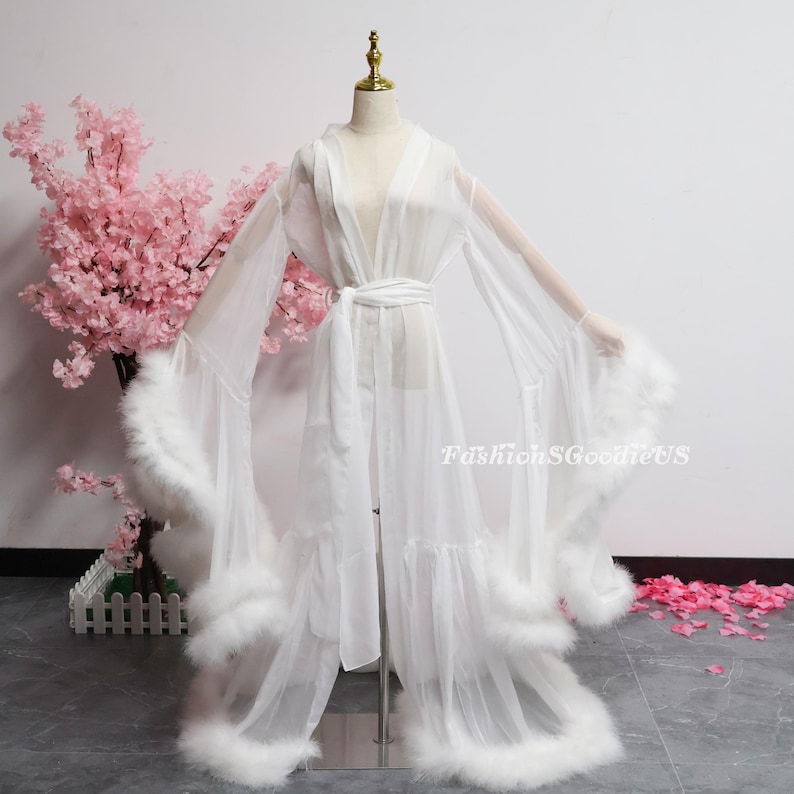 May include: A white, sheer robe with long sleeves and a matching belt. The robe has fluffy white feather trim on the sleeves and hem. The garment is displayed on a mannequin, with a pink floral arrangement in the background.