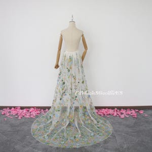 May include: A white tulle skirt with a floral embroidery design. The skirt is on a white mannequin and has a long train.