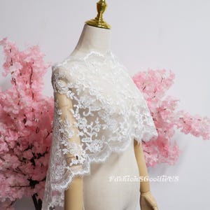 May include: A white lace shawl featuring intricate floral embroidery and scalloped edges draped over a mannequin. The delicate fabric displays a detailed botanical pattern with sheer sections throughout the design. The text FashionSGoodieUS is visible.