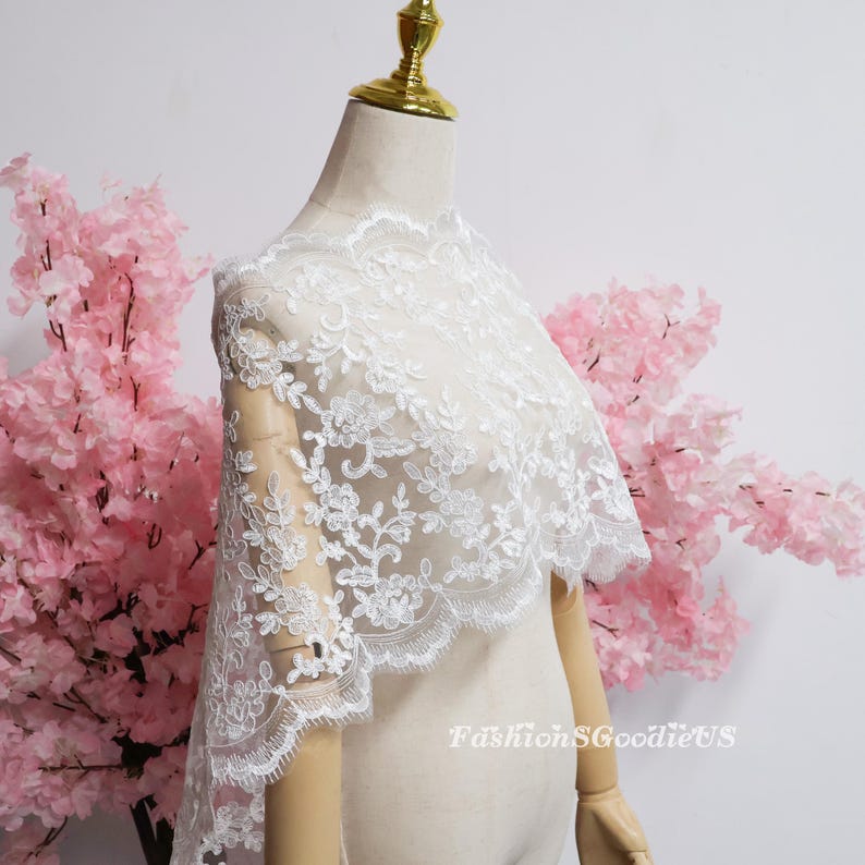 May include: A white lace shawl featuring intricate floral embroidery and scalloped edges draped over a mannequin. The delicate fabric displays a detailed botanical pattern with sheer sections throughout the design. The text FashionSGoodieUS is visible.