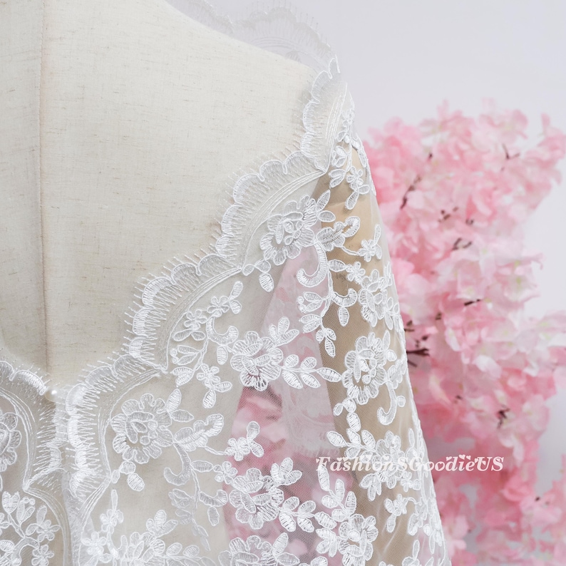 May include: A white lace fabric featuring intricate floral embroidery and scalloped edges draped over a mannequin. The delicate pattern includes detailed leaves and blossoms. The text FashionsGoodieUS is visible in the lower right corner.
