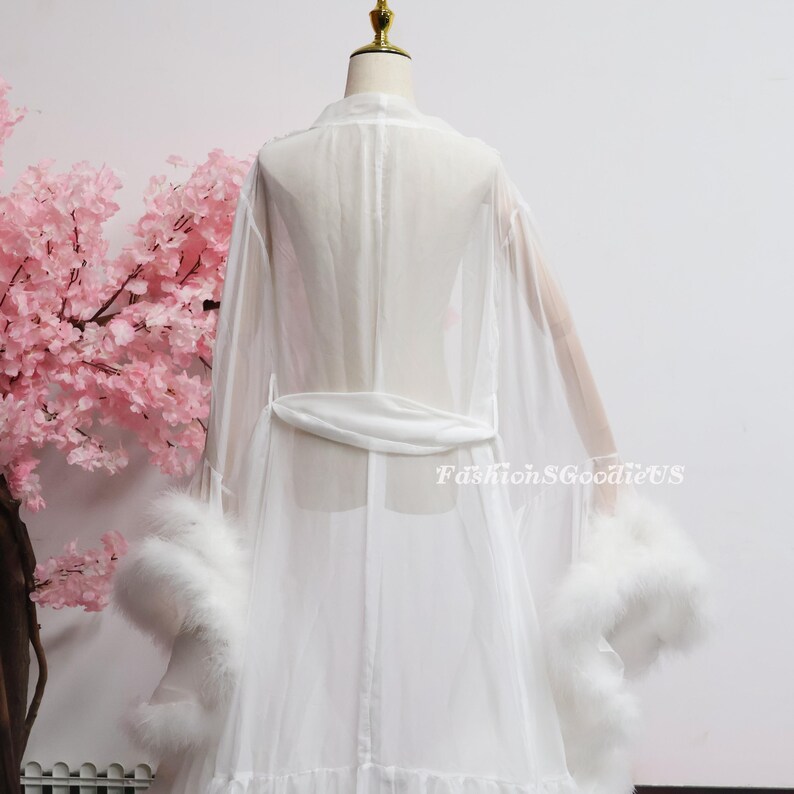 May include: A white, sheer robe with a matching belt and feather trim on the sleeves. The robe is displayed on a mannequin with a pink floral background. The text "FashionGoodieUS" is visible on the lower right side of the image.