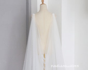 Pode incluir: Uma capa de noiva de tule branco com um delicado adorno de pérolas. A capa está drapeada sobre um manequim e tem as costas baixas.