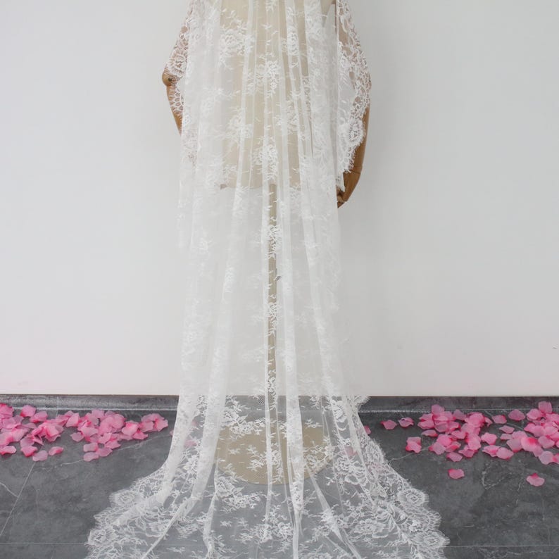 May include: A white lace wedding veil with a delicate floral pattern. The veil is long and flowing, and it is displayed on a mannequin.