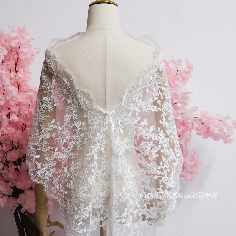 May include: A white lace shawl featuring intricate floral embroidery and a scalloped edge, draped over a mannequin. The sheer fabric displays a delicate pattern of leaves and blossoms. The text FashionSGoodieUS is visible in the lower right corner.