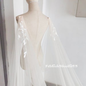 May include: A white tulle bridal cape with delicate floral embroidery. The cape has a sheer, flowing design and is perfect for adding a touch of elegance to any wedding dress.