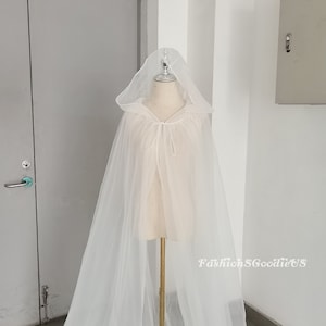 May include: A white tulle cape with a hood. The cape is tied at the neck with a white ribbon. The cape is on a white mannequin.