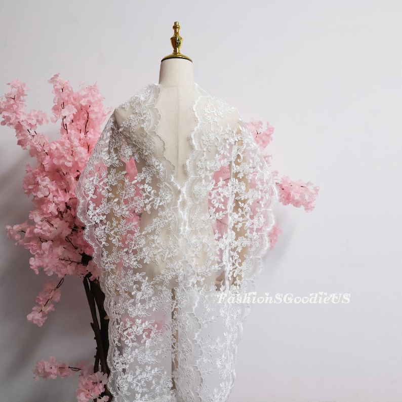May include: A white lace fabric featuring an intricate floral embroidery pattern with scalloped edges. The delicate, semi-sheer material is draped over a mannequin, showcasing the detailed needlework and the text FashionSGoodieUS in the corner.