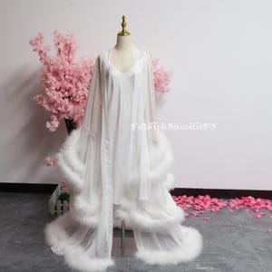 May include: A white lingerie set on a mannequin, comprising a slip dress and a sheer robe. The robe has a long, flowing design with feather trim. The background includes pink cherry blossom branches and the text "FashionSGGoodieUS".