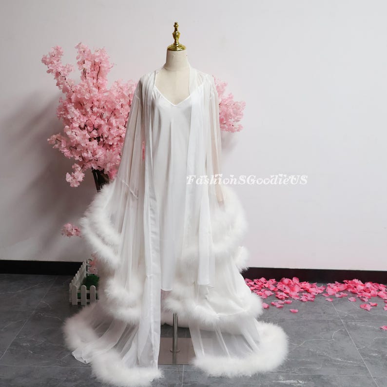 May include: A white lingerie set on a mannequin, comprising a slip dress and a sheer robe. The robe has a long, flowing design with feather trim. The background includes pink cherry blossom branches and the text "FashionSGGoodieUS".