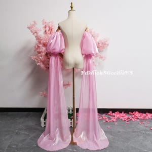 May include: A light pink off-the-shoulder garment with voluminous sleeves and long, flowing fabric extensions. The garment is displayed on a mannequin, with the text "FashionSGoodieUS" visible. The background features pink floral arrangements.
