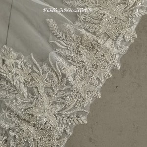May include: Close-up of a white lace fabric with intricate floral designs and sparkling embellishments. The fabric is sheer and delicate, with a soft, flowing texture.