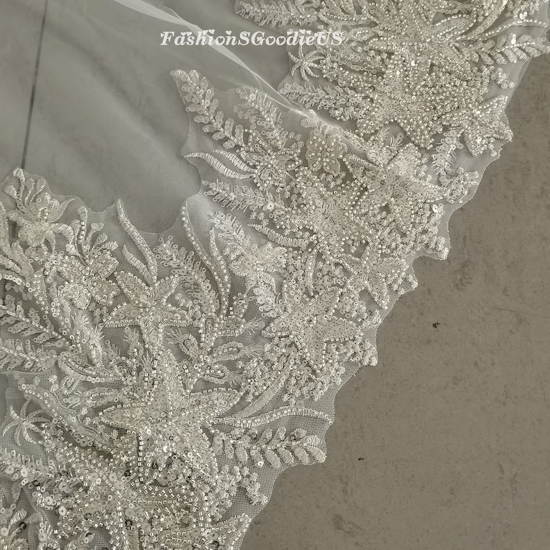 May include: Close-up of a white lace fabric with intricate floral designs and sparkling embellishments. The fabric is sheer and delicate, with a soft, flowing texture.