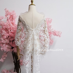 May include: A white lace shawl with an intricate floral pattern and scalloped edges draped over a mannequin. The delicate fabric features a sheer mesh base with detailed embroidery. The text FashionSGoodieUS is visible on the lower right side.