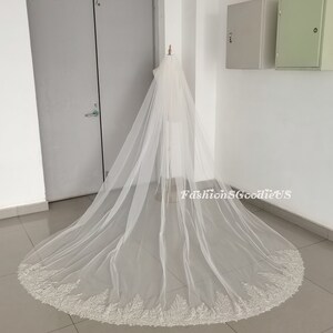 May include: A long, white bridal veil with a beaded lace trim. The veil is draped over a mannequin and is shown against a white background.