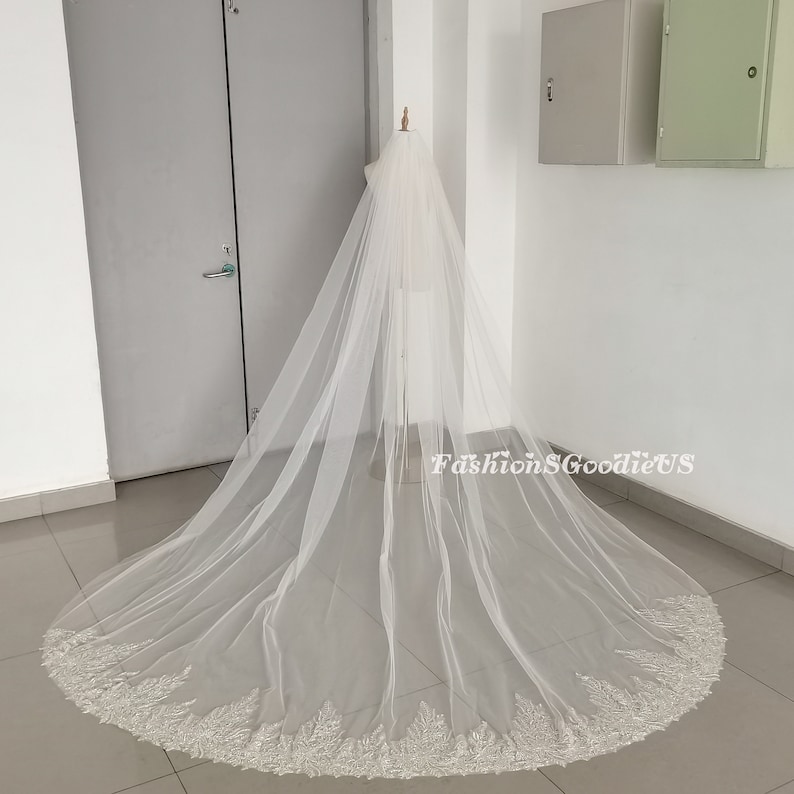 May include: A long, white bridal veil with a beaded lace trim. The veil is draped over a mannequin and is shown against a white background.