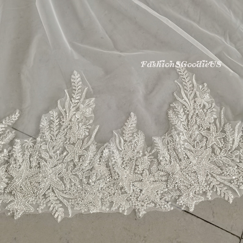 May include: White tulle wedding veil with a beaded floral lace trim. The lace features a delicate floral design with small, sparkling beads.