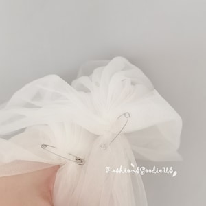 May include: White tulle fabric is gathered and secured with two safety pins. The fabric is bunched up and ready to be attached to a headpiece.