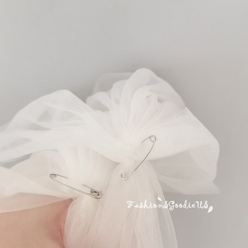 May include: White tulle fabric is gathered and secured with two safety pins. The fabric is bunched up and ready to be attached to a headpiece.