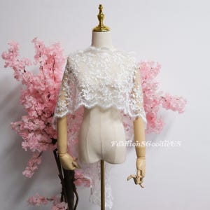 May include: A white lace bolero shrug featuring an intricate floral embroidery pattern with scalloped edges. The garment is displayed on a mannequin and includes the text FashionSGoodieUS printed in white on the lower right side of the lace fabric.