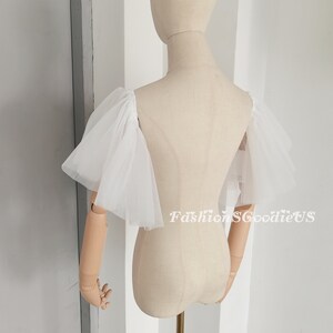 Pu&ograve; includere: Maniche in tulle bianco con un dettaglio di volant arricciati. Le maniche sono mostrate su un manichino bianco.