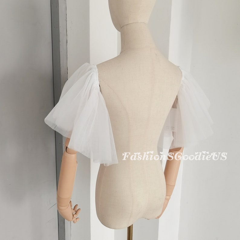 Pu&ograve; includere: Maniche in tulle bianco con un dettaglio di volant arricciati. Le maniche sono mostrate su un manichino bianco.
