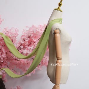 May include: A light green scarf draped over a mannequin. The scarf is made of a lightweight fabric and has long, flowing ends. The mannequin is positioned in front of pink cherry blossom branches. The text "FashionSGoodieUS" is visible on the mannequin.