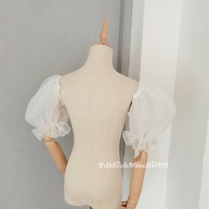 May include: White, sheer, puff sleeves with ruffled cuffs. The sleeves are displayed on a mannequin. The sleeves are made of a lightweight, airy fabric. The overall style is romantic and delicate.