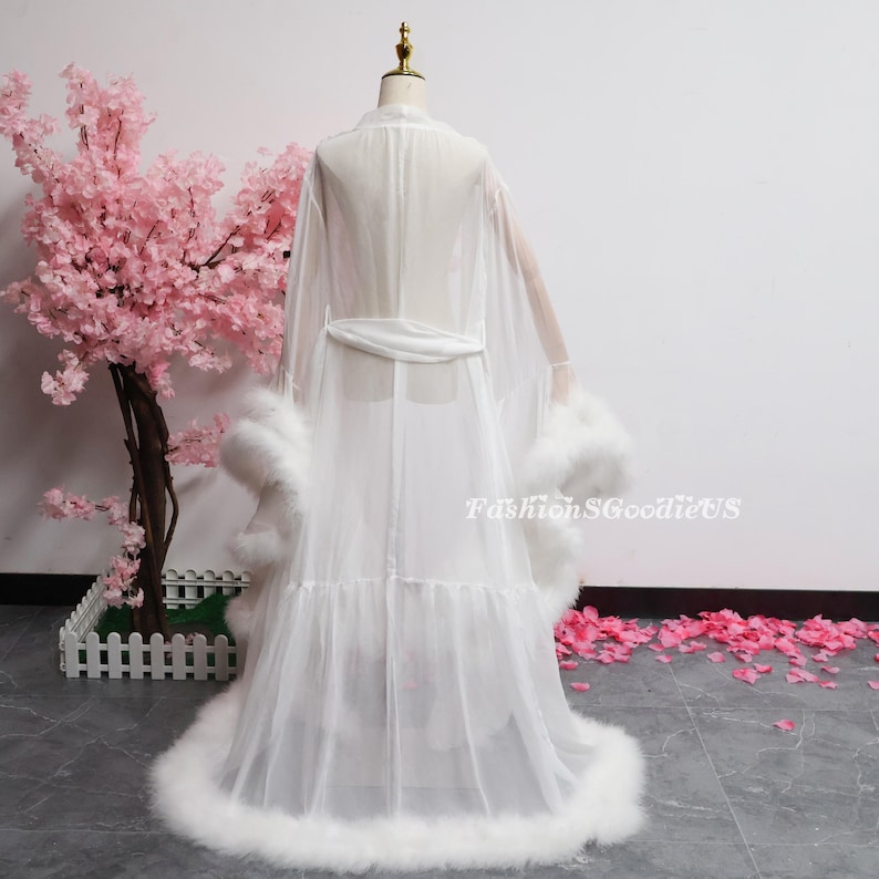 May include: A white, sheer robe with feather trim on the cuffs and hem, displayed on a mannequin. The robe has long sleeves and a matching belt. The text "FashionSGoodieUS" is visible on the lower right.