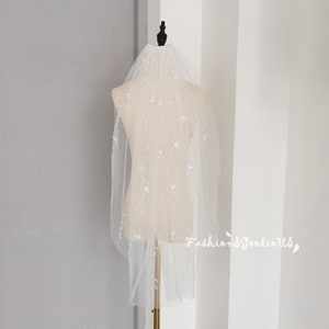 May include: A white bridal veil with a delicate floral pattern and scattered rhinestones. The veil is displayed on a mannequin.