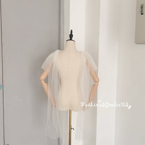 May include: A white tulle cape with a ruffled neckline and long sleeves. The cape is draped over a white mannequin.