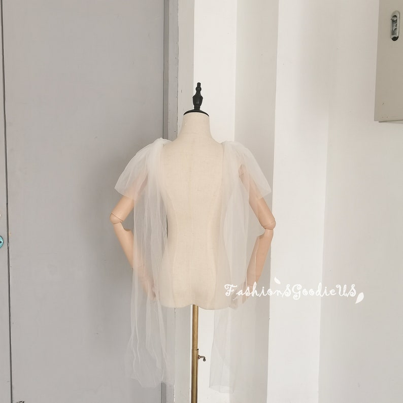 May include: A white tulle cape with a ruffled neckline and long sleeves. The cape is draped over a white mannequin.