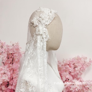 May include: A white bridal veil with intricate lace detailing and floral embellishments. The veil is draped over a mannequin head, showcasing its delicate design and texture. Pearls are scattered throughout the veil.