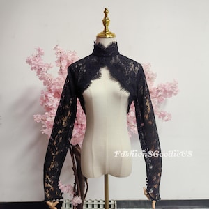 May include: Black lace bolero with long sleeves and a high neckline. The bolero is displayed on a mannequin with a gold-coloured head. The sleeves have small, decorative buttons at the wrist. The background includes pink flowers.