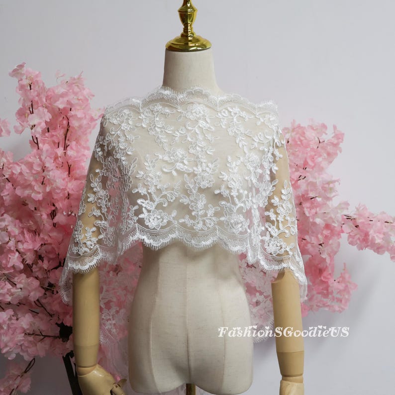 May include: A white lace shawl featuring intricate floral embroidery and a scalloped edge, draped over a mannequin. The delicate fabric displays a detailed botanical pattern throughout, with the text FashionSGoodieUS visible in the lower right.
