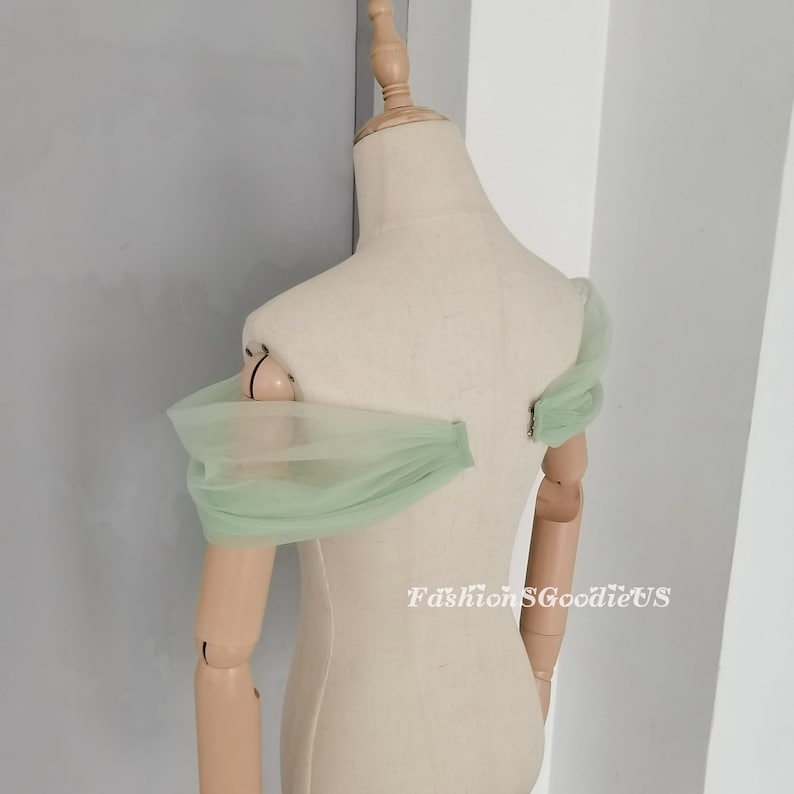 May include: A pair of light green tulle off-the-shoulder sleeves. The sleeves are gathered at the shoulders and have a soft, flowing drape.
