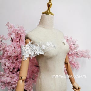May include: A white floral lace sleeve is displayed on a mannequin. The delicate lace features intricate flower and leaf patterns. The sleeve is designed to be worn off the shoulder. The mannequin is positioned in front of pink cherry blossom branches.